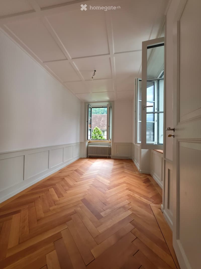 Empty room with wooden herringbone flooring, white walls, and large windows letting in natural light. The room has a radiator and a door leading to another space.