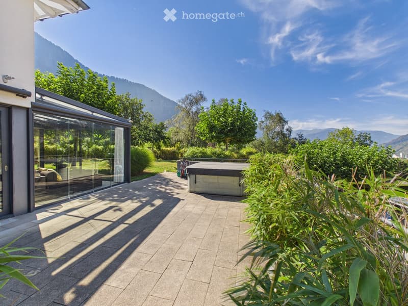 A modern outdoor patio with a tiled floor, glass enclosure, and a hot tub, surrounded by lush greenery and a scenic mountain view.