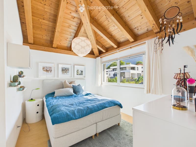 Cozy bedroom with wooden slanted ceiling, large window with view, bed with blue duvet, nightstand with lamp, decorative items including a dreamcatcher and toy ship.