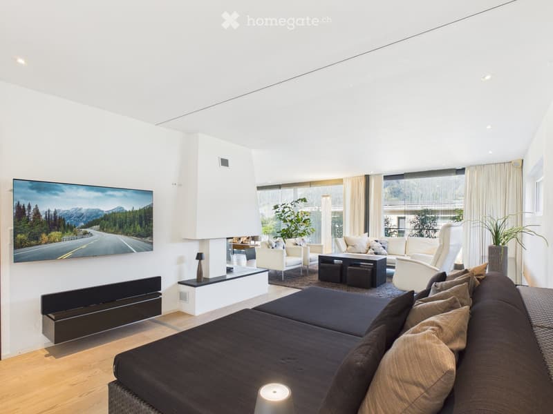 Modern living room with large windows, white walls, and a mix of seating including a dark sectional sofa, white armchairs, and a coffee table. A flat-screen TV is mounted on the wall, and there is a fireplace with a decorative screen. The room features a
