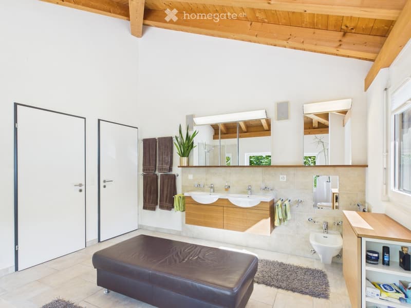 Modern bathroom with wooden cabinets, double sink, large mirror, leather stool, and towels.