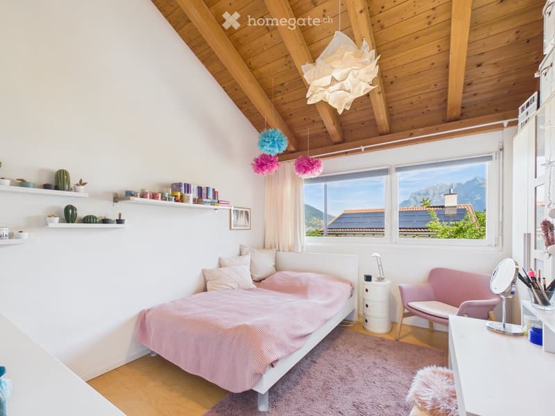 Bright bedroom with wooden ceiling beams, large windows overlooking mountains, white bed with pink bedding, pink sofa, shelves with books and decorative items, and a vanity area with a mirror.