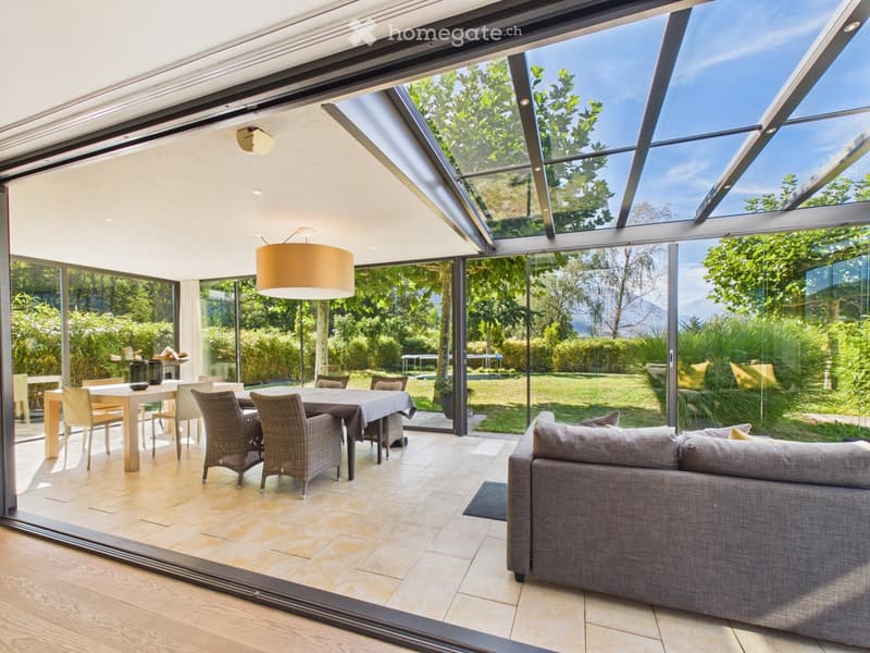Bright, modern living and dining area with large glass walls and a skylight, featuring a grey sofa, dining table with chairs, and a hanging lamp.