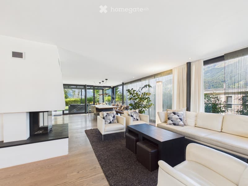 Modern living room with large windows offering a scenic view, featuring a white sofa, coffee table, armchairs, ottomans, and a fireplace.