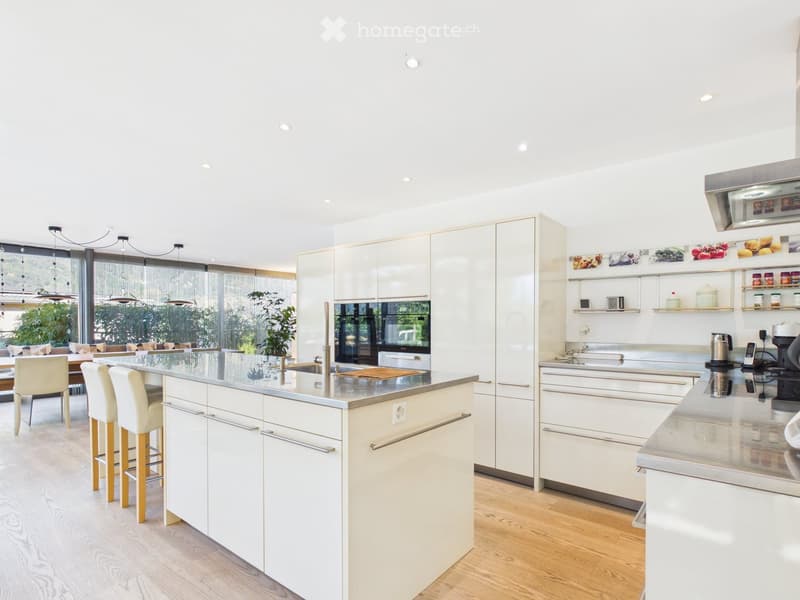 Modern kitchen with white cabinets, a central island, and a dining area visible through large windows. The island features a sleek countertop and four white stools. The kitchen includes built-in appliances, open shelving, and a dining area with a table an