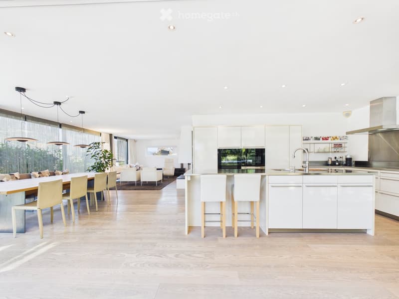 Open-plan kitchen and dining area with white cabinets, a central island, pendant lights, and a dining table with chairs. The space features light wood flooring and large windows with sheer curtains.