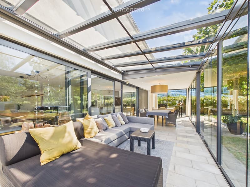 Bright, modern living space with large glass walls and a retractable roof, featuring a grey sectional sofa with yellow cushions, a small coffee table, and a dining area with a table and chairs.
