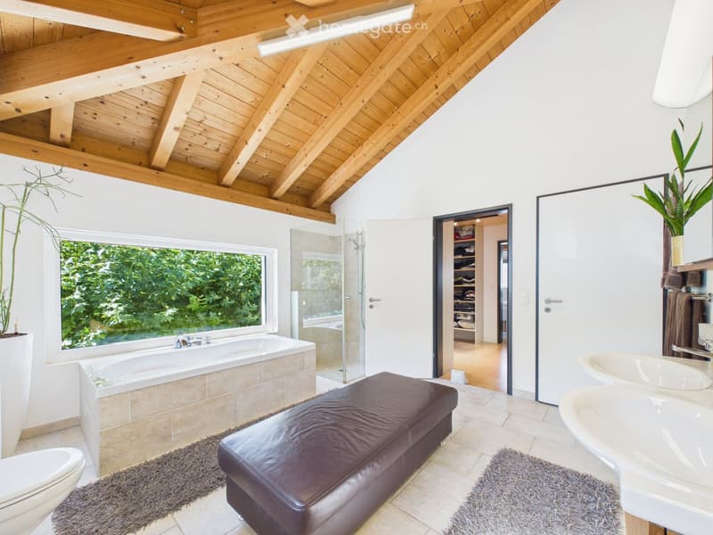Modern bathroom with a large window offering a view, featuring a bathtub, toilet, double sink, leather stool, and a glass-enclosed shower area.