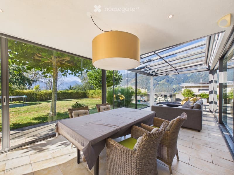 Bright, airy room with large glass walls offering a scenic mountain view. Features a dining table with chairs, a sofa, and a hanging lamp.