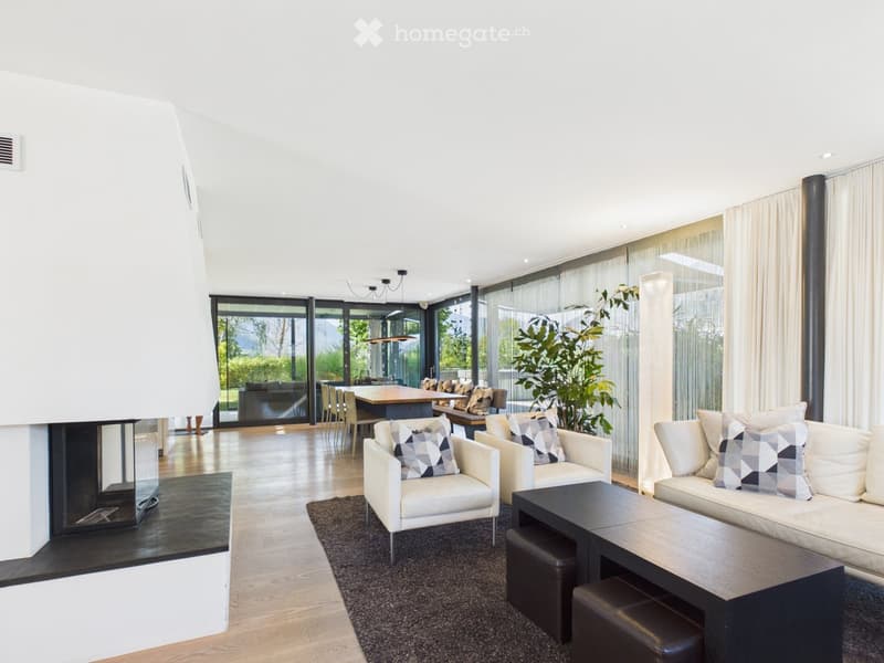 Modern living room with white walls, wooden flooring, large windows, fireplace, sofa, coffee table, chairs, ottomans, and a dining area visible through the glass doors.