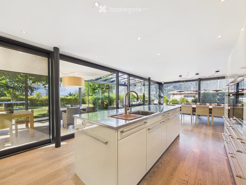 Modern kitchen with white cabinets, stainless steel sink, and large windows overlooking a garden. Includes a dining area with chairs and a table.