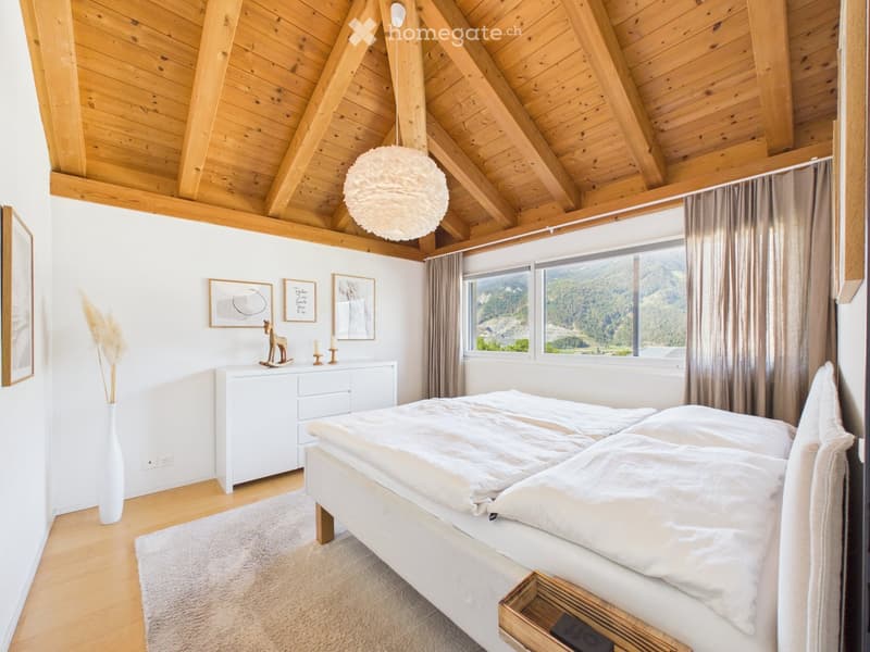 Cozy bedroom with wooden ceiling beams, large windows offering a mountain view, a neatly made bed, a white nightstand, a decorative vase, and framed artwork on the walls.