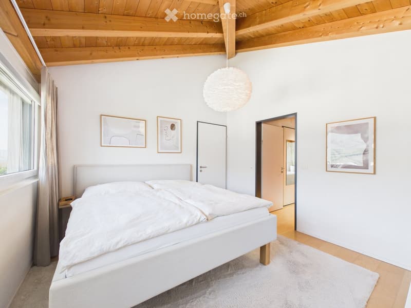 Bright bedroom with a white bed, nightstand, pendant light, and artwork on the walls. The room features wooden flooring and a slanted wooden ceiling.