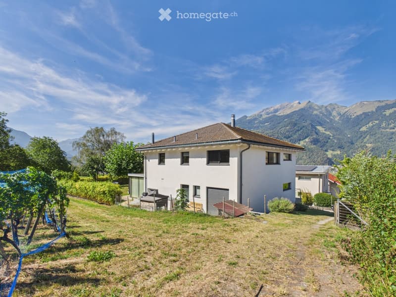 Two-story white house with a brown roof, surrounded by a grassy yard, trees, and mountains in the background.