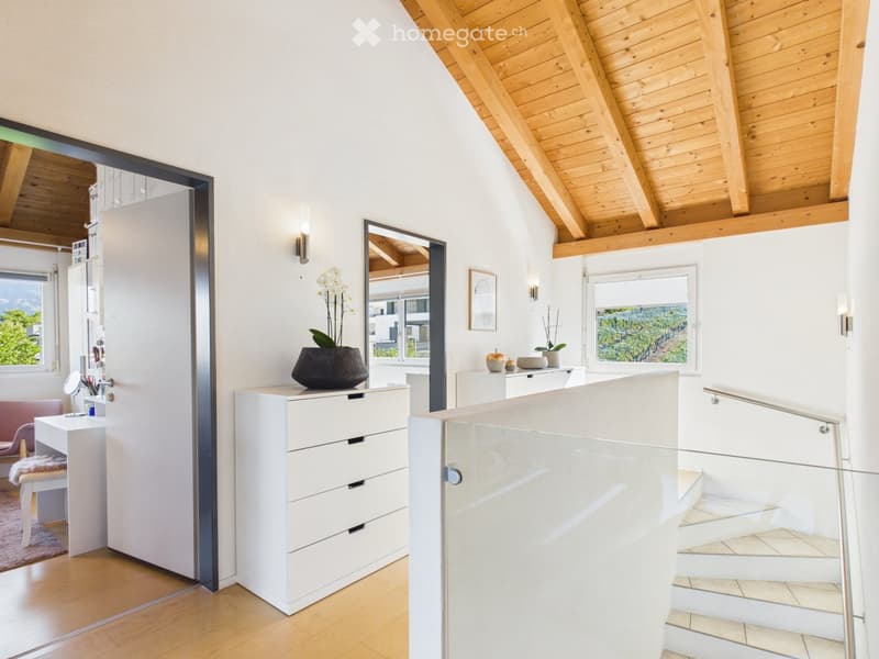 Modern, well-lit room with wooden floors, white walls, and a slanted wooden ceiling. Features include a vanity with a mirror, a small table with a vase, a drawer unit, and a staircase with a glass railing.