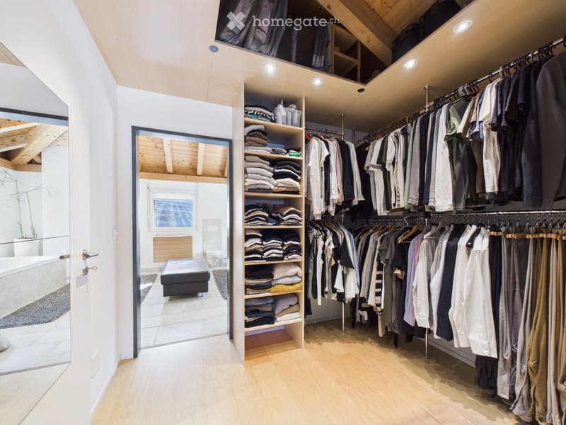 Spacious walk-in closet with wooden flooring, built-in wardrobe, shelves, and an adjacent bathroom with a bathtub and toilet.