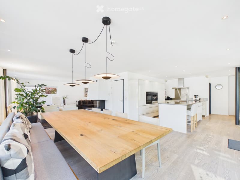 Open-plan living and dining area with a large wooden dining table, white kitchen island, modern pendant lights, and a comfortable sofa.