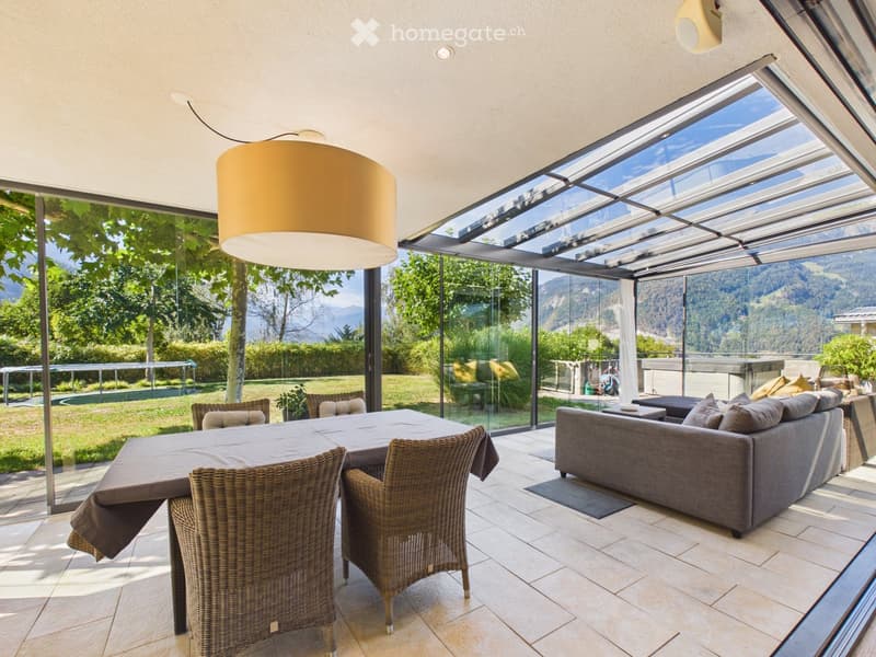 Bright, modern living space with large glass walls offering scenic mountain views, featuring a sofa, dining table, and wicker chairs.