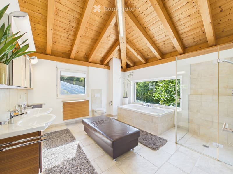 Modern bathroom with wooden ceiling beams, large windows offering a view, double sinks, a bathtub, a toilet, a leather stool, and a glass-enclosed shower.