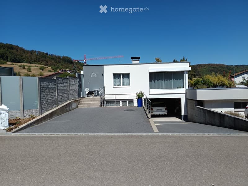 Modern two-story house with white exterior, garage, and outdoor seating area. The house features large windows, a stone wall, and a paved driveway.