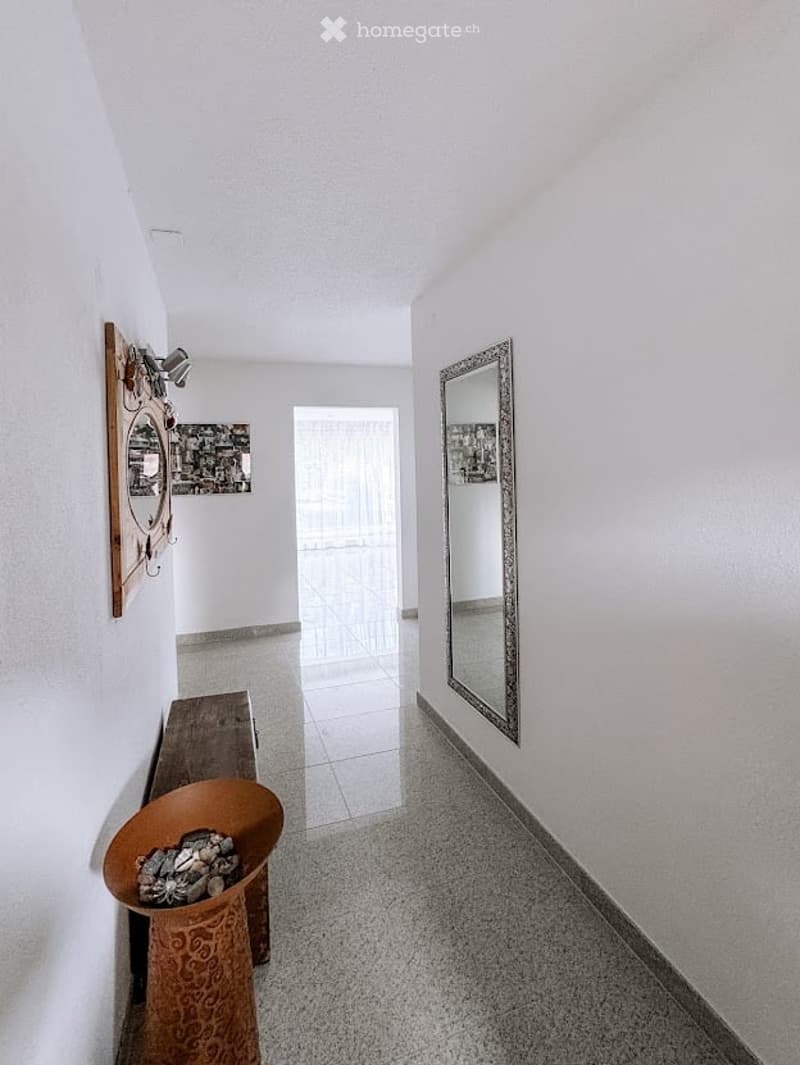 A clean, modern hallway with white walls, tiled flooring, and a large mirror on one side. A small wooden bench and a decorative bowl are placed near the wall.