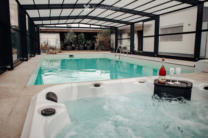 Indoor pool area with a swimming pool, a hot tub, and a bottle of wine with two glasses on a table.