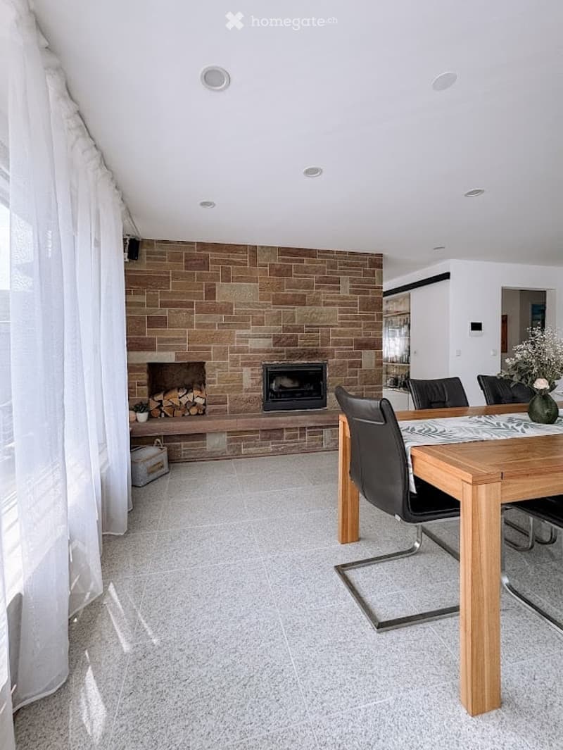 Modern dining area with a fireplace, wooden dining table, black chairs, and a large window with white curtains.