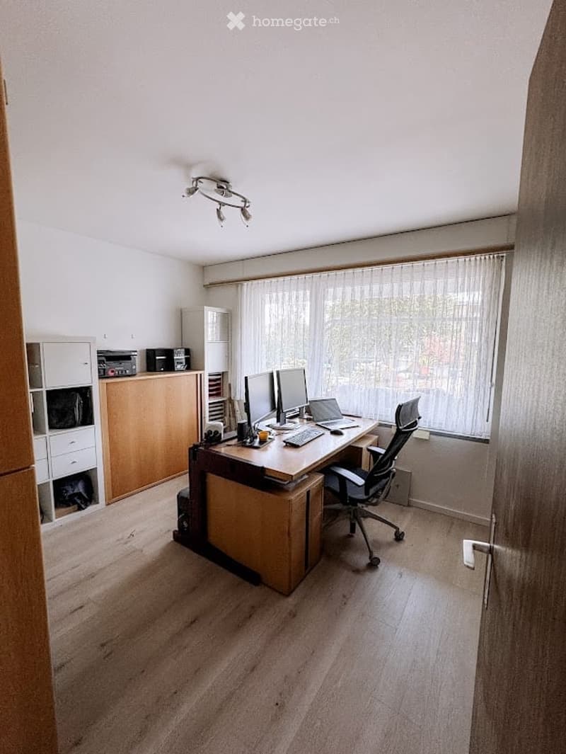A well-lit office space with a large wooden desk, ergonomic chair, dual monitors, laptop, keyboard, mouse, and storage cabinets.