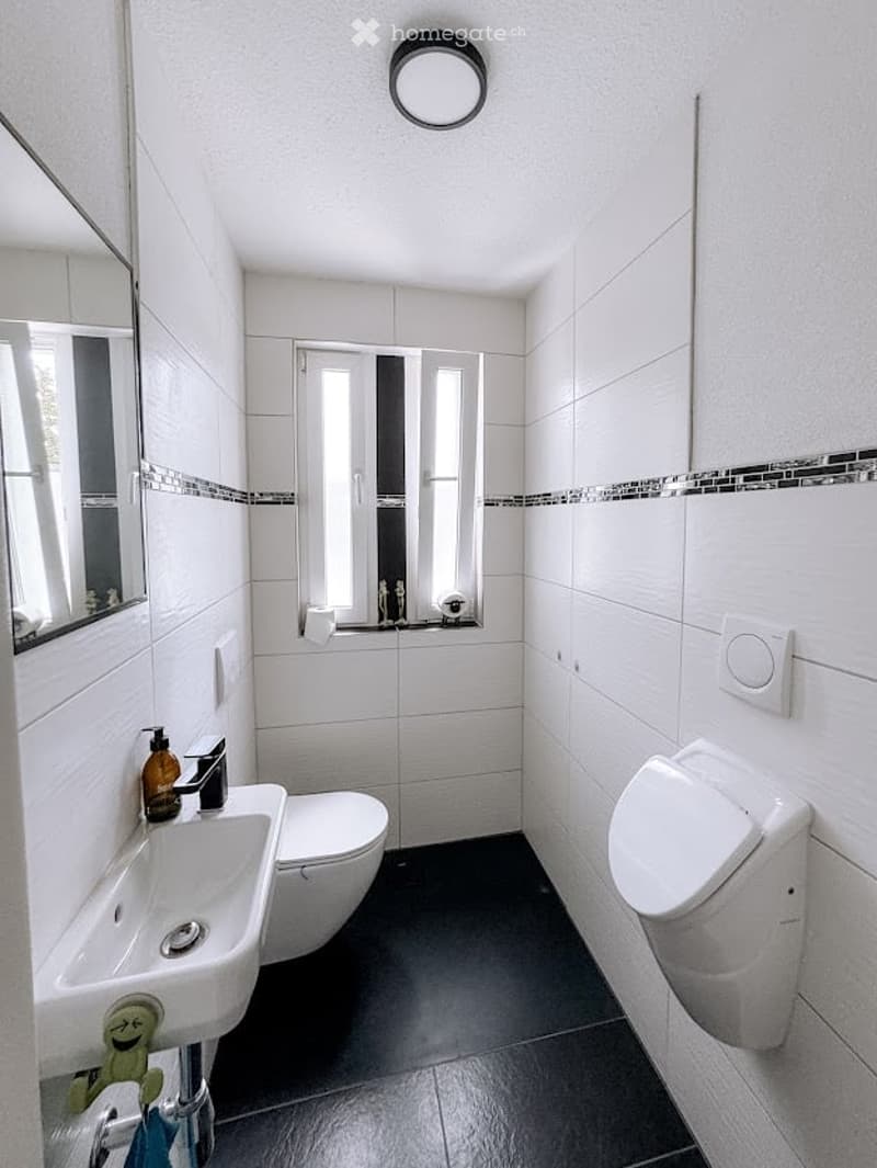 Modern bathroom with white tiles, a sink, toilet, mirror, and a soap dispenser. The floor is dark grey, and there is a small window above the toilet.