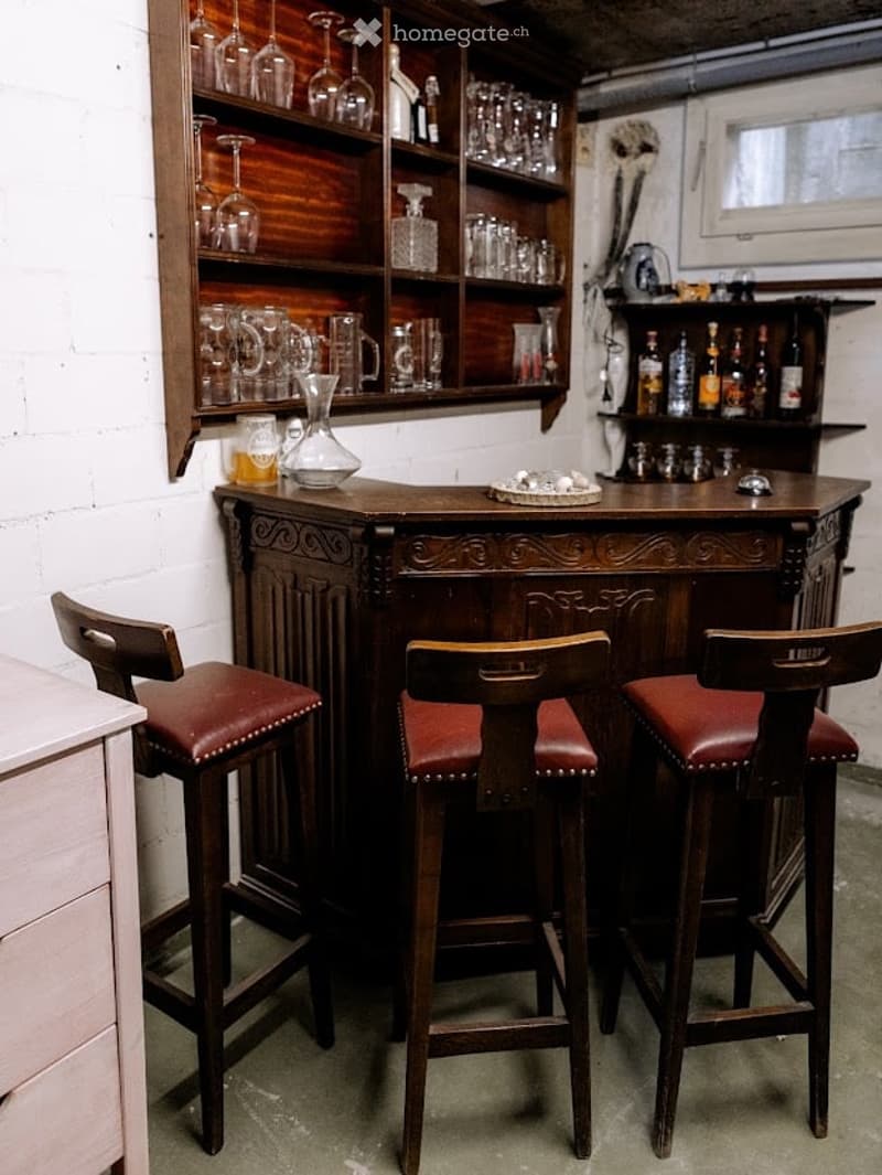 A cozy bar area with a wooden bar counter, three leather-topped bar stools, and shelves filled with glassware and bottles.