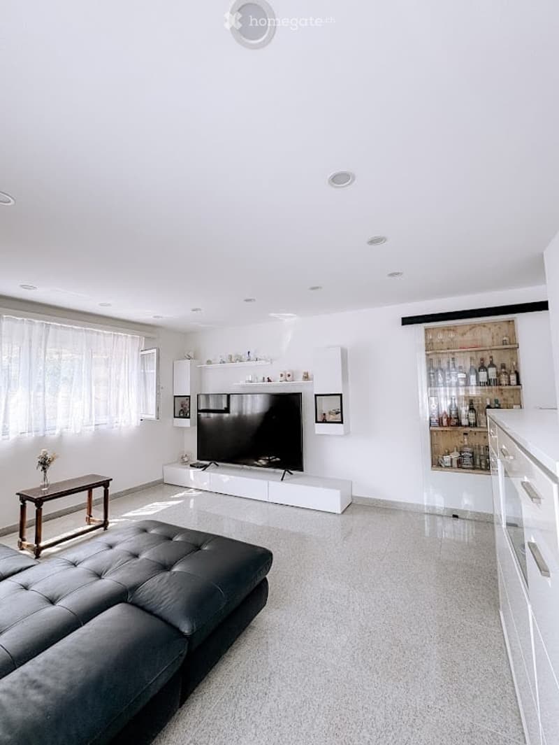 Modern living room with white walls, grey carpet, large windows, a black leather sofa, a wooden coffee table, a flat-screen TV on a white cabinet, and a glass cabinet with bottles and decorative items.