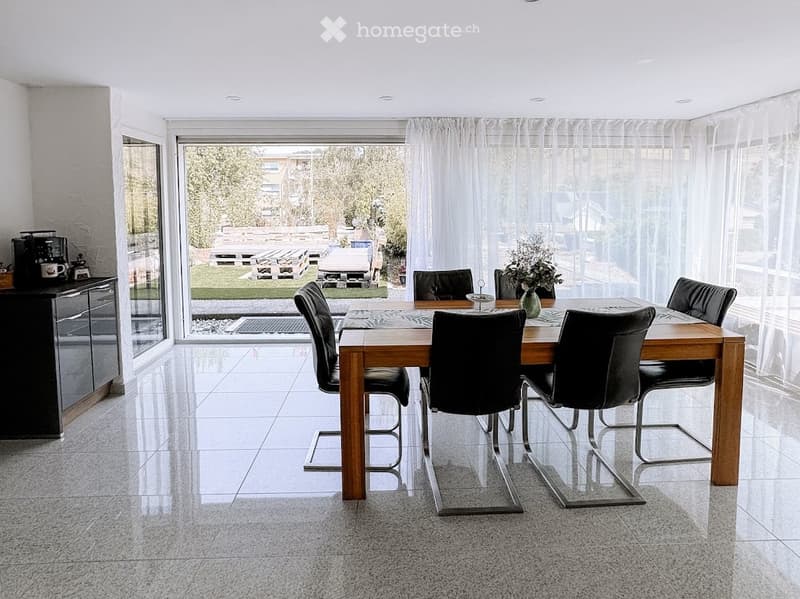 Modern dining area with a wooden dining table, black chairs, a sleek black coffee machine on a cabinet, and a vase with flowers on the table. Large windows offer a view of the garden.