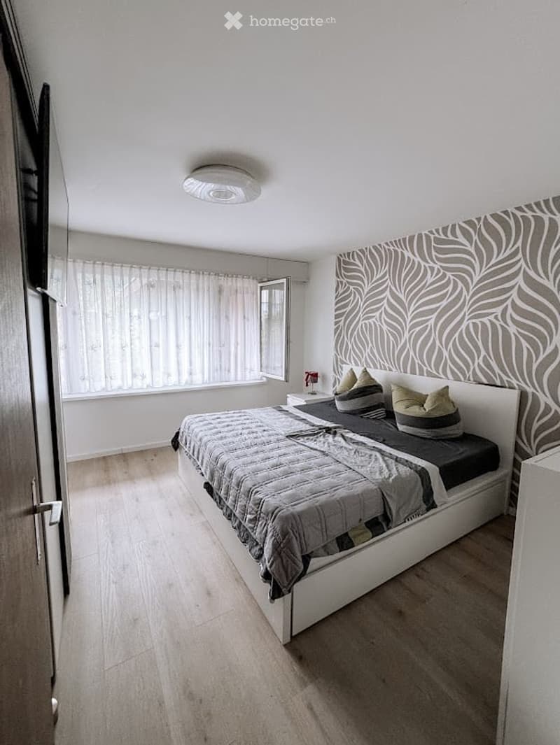 Modern bedroom with a large bed, nightstand, and decorative pillows. The room features light wooden flooring, white walls, and a patterned accent wall. A window with sheer curtains allows natural light to enter.