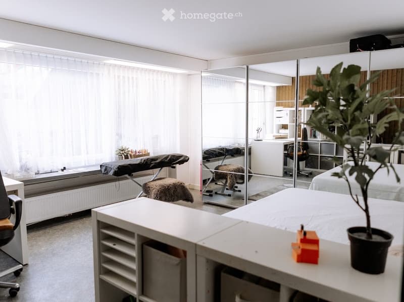 Bright, minimalist room with a large mirror, a small table with a plant, a chair, and a stool. The room features a white color scheme with a modern aesthetic.