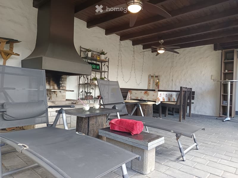 Outdoor patio with a stone table, chairs, a bench, and a small shelf with plants. A ceiling fan is visible, and the area is covered with a wooden ceiling.