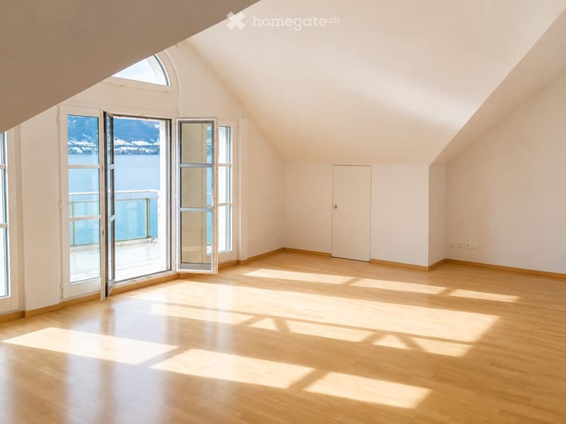 Bright, empty room with wooden flooring and large windows offering a scenic view. The space features a door and ample natural light.