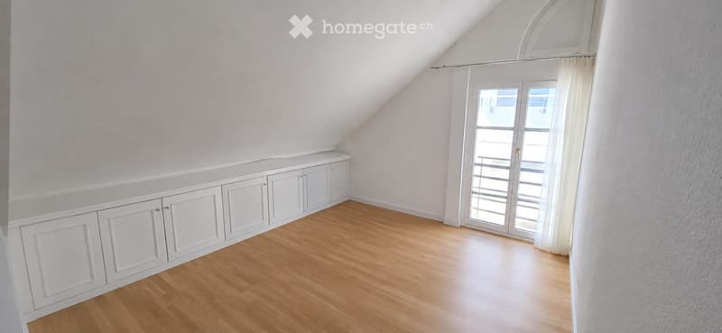 Empty attic room with wooden flooring, white built-in cabinets along one wall, and a large window with white curtains.