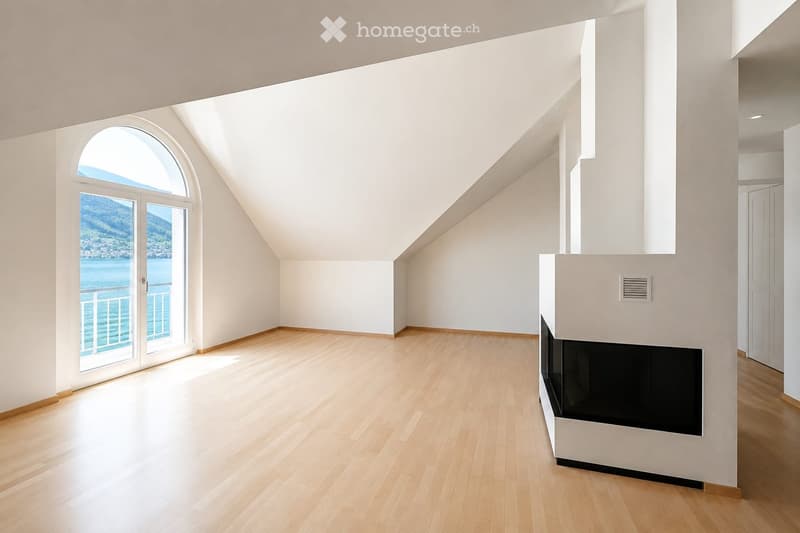 Bright, empty room with wooden flooring, white walls, and a fireplace. Large arched window offers a scenic view of the water and mountains.