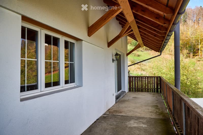 Wooden balcony with a wooden railing, overlooking a green landscape with trees and hills.