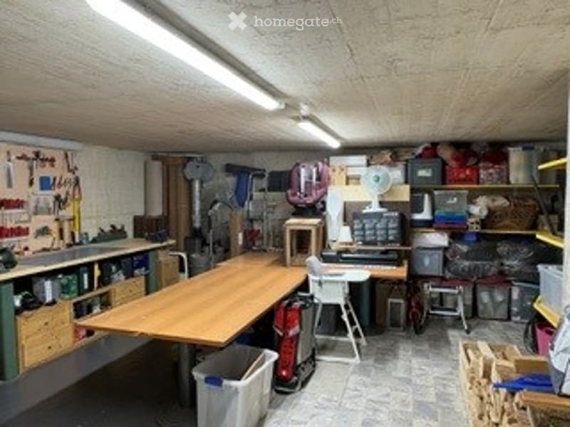 A well-organized workshop or garage space with a large wooden worktable, various tools on the walls, storage bins, a computer setup, and a small refrigerator.
