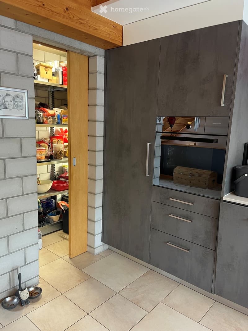 Modern kitchen with grey cabinets, an oven, and a pantry area with shelves holding various items. The floor is tiled, and there is a pet bowl and toy on the floor near the wall.