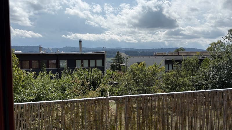 Balcony with wooden railing overlooking a landscaped area with trees and two modern buildings in the background, under a partly cloudy sky.