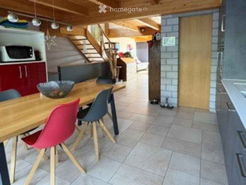 Bright, modern dining area with a wooden dining table, colorful chairs, and a microwave in the background. The space features exposed wooden beams and a staircase leading to another level.