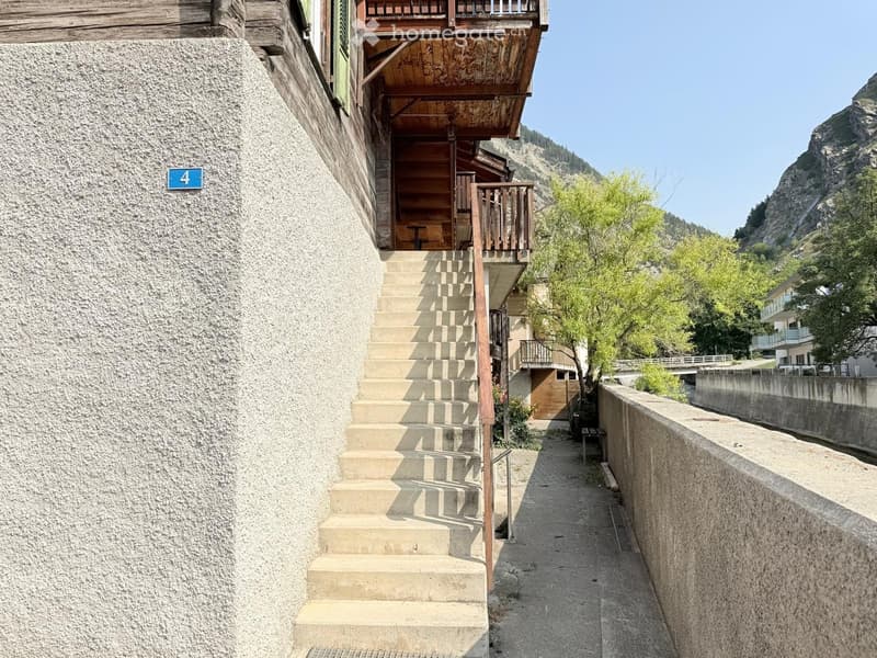 Narrow stone-paved pathway leading to a wooden staircase and balcony, with a stone wall on one side and a wooden building on the other.