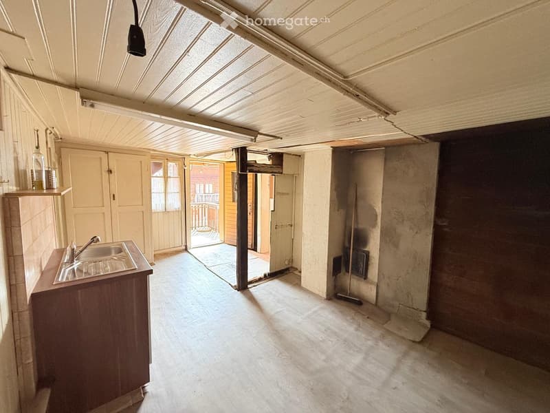 Empty room with wooden flooring, exposed ceiling beams, and a small sink area. The space has large windows and doors leading to an outdoor area.