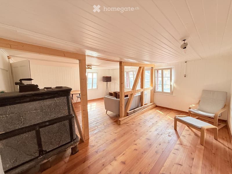 Bright, wooden-floored room with exposed wooden beams, a fireplace, a sofa, chairs, a small table, and a floor lamp.