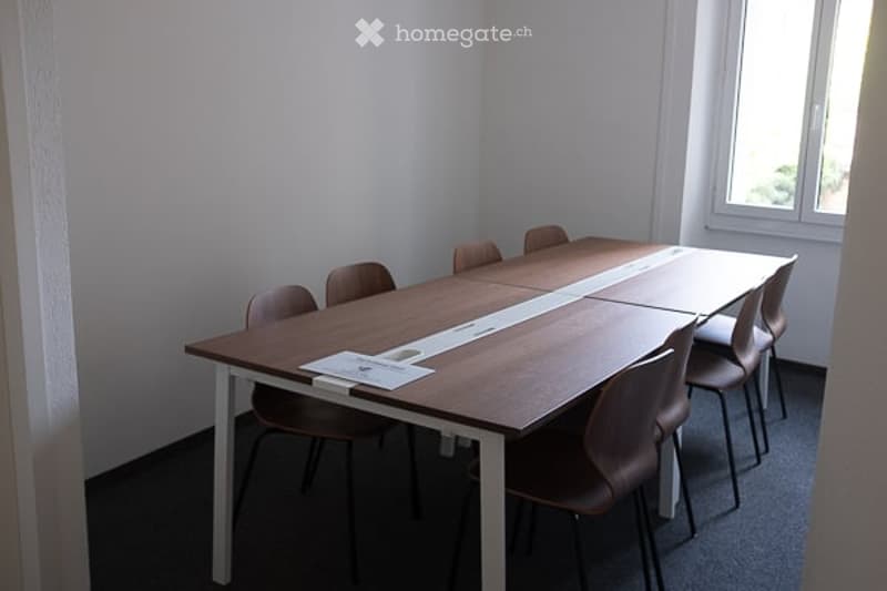 A small, modern meeting or conference room with a long wooden table and several chairs arranged around it. The room has white walls, a large window, and a carpeted floor.
