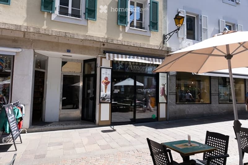 Outdoor seating area with a table, chairs, and an umbrella in front of a building with a storefront and green shutters.