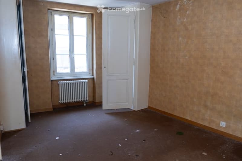 Empty room with bare walls, wooden floor, single window, white door, and radiator.