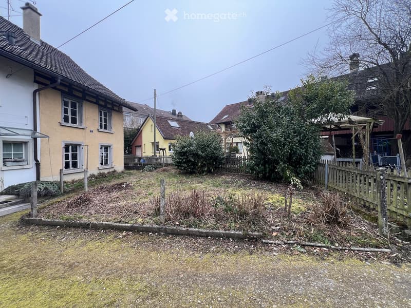 A small, fenced garden area with sparse vegetation and a wooden fence, surrounded by neighboring houses with varied architectural styles and colors.